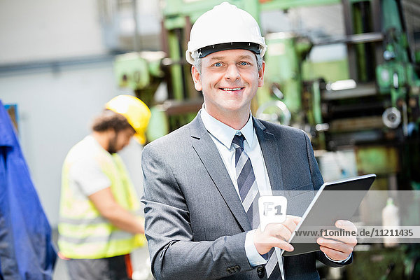 Porträt eines selbstbewussten reifen Geschäftsmannes  der ein digitales Tablet benutzt  mit einem Arbeiter im Hintergrund in einer Fabrik