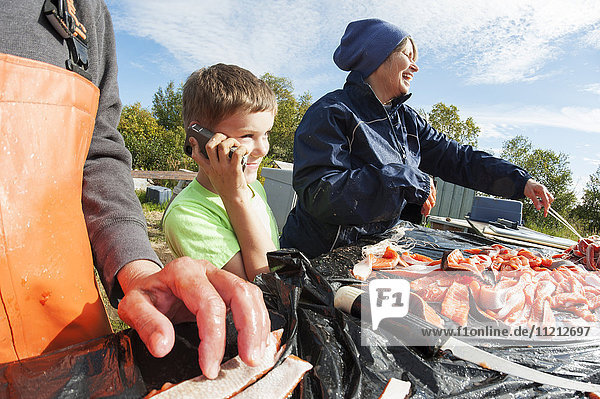 Young boy talks on a cell phone while adults filet and hang caught salmon Bristol Bay Alaska