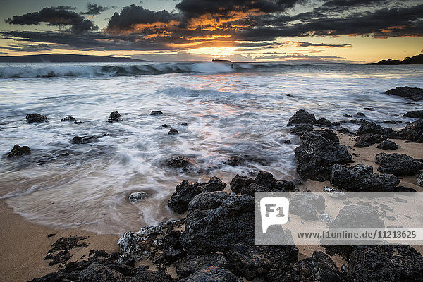 Wasser  das auf die schwarzen Felsen und den Sand am Strand bei Sonnenuntergang gespült wird; Maui  Hawaii  Vereinigte Staaten von Amerika'.
