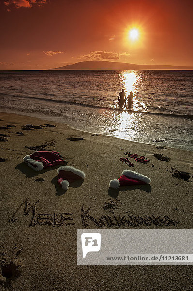 Paar  das bei Sonnenuntergang im Meer watet  mit Weihnachtsschmuck und dem Schriftzug Mele Kalikimaka im Sand; Maui  Hawaii  Vereinigte Staaten von Amerika'.