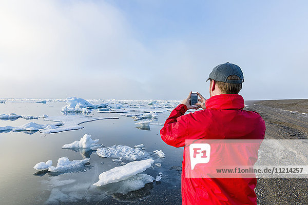 A man standing on the coast of the Arctic Ocean during a foggy sunset takes a picture with his cell phone with sea ice floating in the background; Barrow; North Slope; Arctic Alaska; USA; Summer