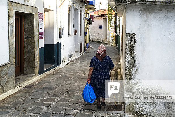Eine ältere Frau geht mit einem Stock und einer blauen Tasche eine schmale Straße zwischen Häusern auf einer griechischen Insel entlang; Glossa  Skiathos  Griechenland'.