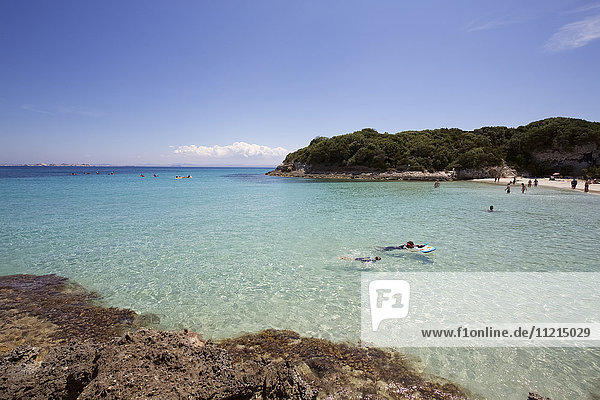 Swimmers bathing in turquoise water of white sand bay bordered by rocky wooded promontory