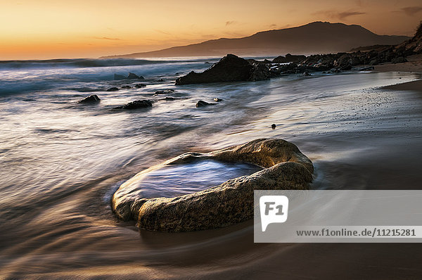 'Water washes up onto the sand and a rock on the beach with an orange sky glowing at sunset; Tarifa  Cadiz  Andalusia  Spain'