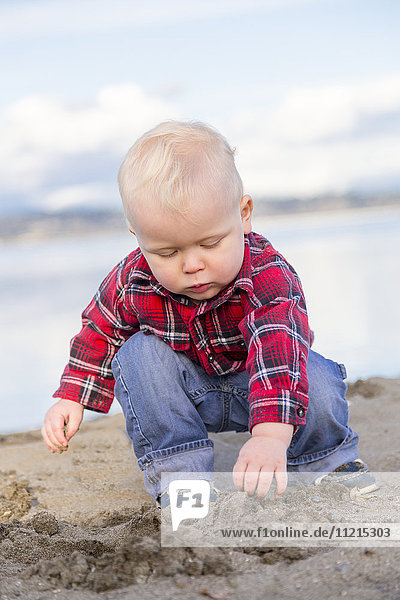 Ein kleiner Junge in einem rotkarierten Hemd und Jeans spielt im Sand am Meer; Surrey  British Columbia  Kanada