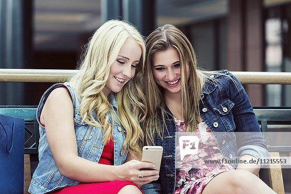 'Two beautiful young women out shopping and taking a break to check out social media on a smart phone; Edmonton  Alberta  Canada'