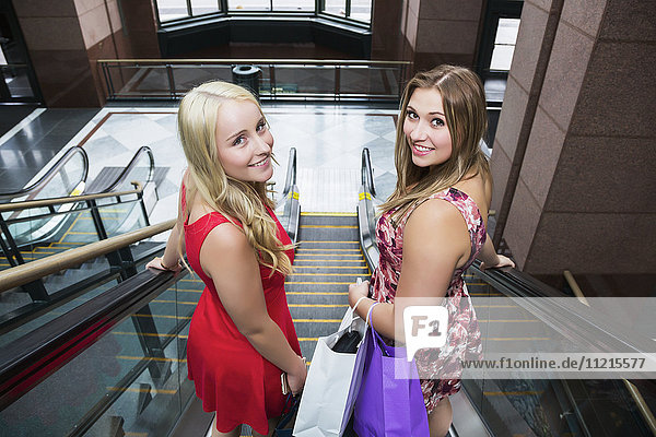 'Two beautiful young women wearing dresses out shopping together and riding an escalator in an indoor mall complex and looking back at camera; Edmonton  Alberta  Canada'