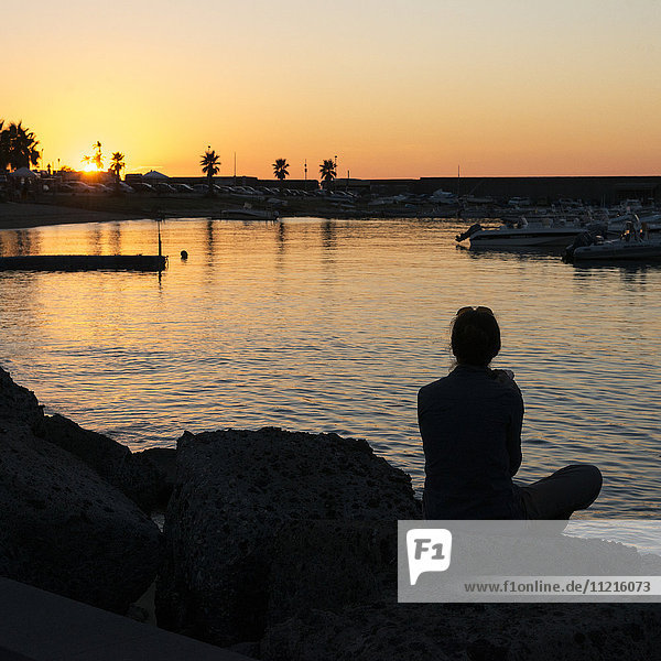 'Silhouette of a woman sitting at the water's edge looking out over a harbour at sunset; Forio  Ischia  Campania  Italy'
