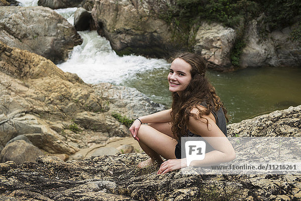 'A teenage girl sitting on a rock posing for the camera with a waterfall in the background; Yalepa  Jalisco  Mexico'