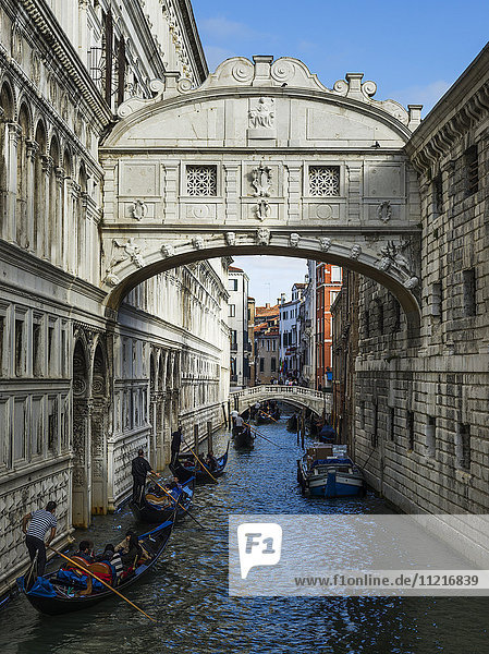 Dekorativer Torbogen über einem Kanal  der zwei Gebäude miteinander verbindet  mit Gondeln und Gondolieri  die den Kanal hinunterpaddeln; Venedig  Italien