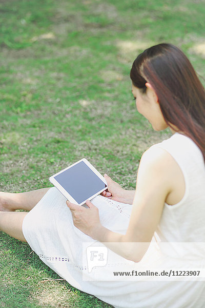 Porträt einer jungen japanischen Frau  die mit einem Tablet im Gras liegt