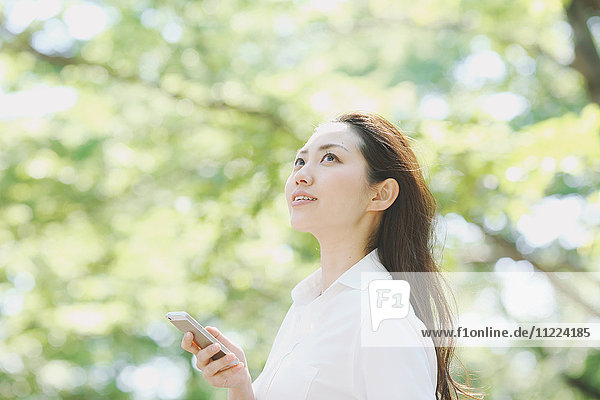 Junge Japanerin mit Smartphone umgeben von Grün in einem Stadtpark