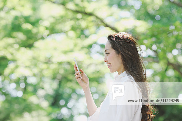 Junge Japanerin mit Smartphone umgeben von Grün in einem Stadtpark