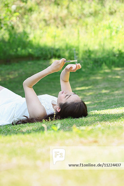 Porträt einer jungen japanischen Frau  die mit ihrem Smartphone im Gras liegt