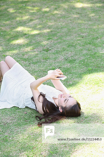 Porträt einer jungen japanischen Frau  die mit ihrem Smartphone im Gras liegt