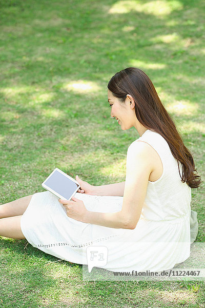 Porträt einer jungen japanischen Frau  die mit einem Tablet im Gras liegt