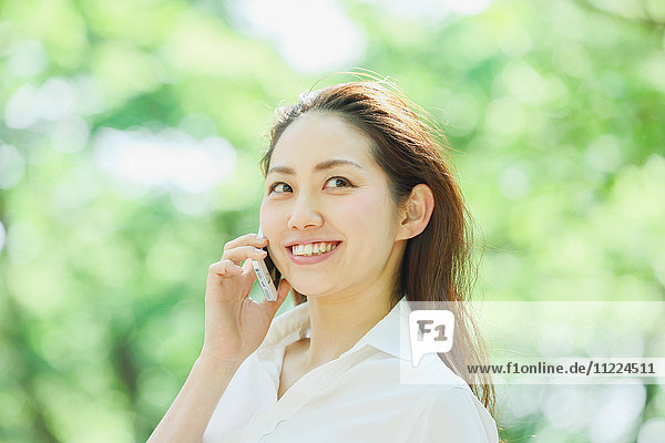 Junge japanische Frau am Telefon  umgeben von Grün in einem Stadtpark