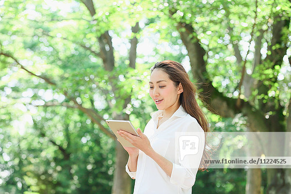 Junge Japanerin mit Tablet inmitten von Grün in einem Stadtpark