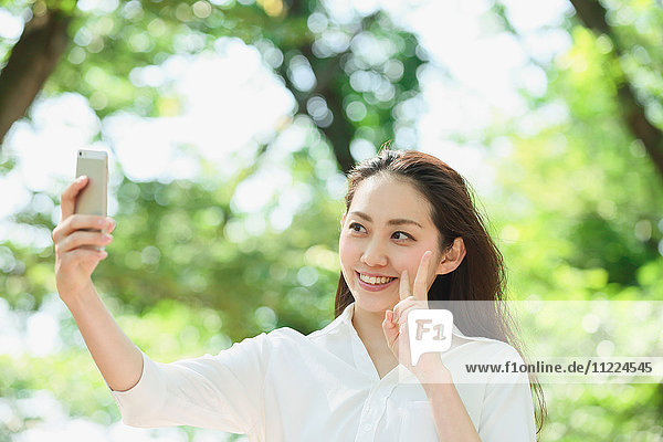 Junge japanische Frau nimmt Selfie umgeben von Grün in einem Stadtpark