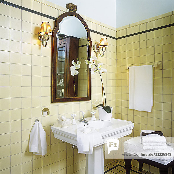 1920s style. Detail of white stately pedestal sink in center and mahogany framed mirror. White orchids sitting on right side of sink. Shade sconces. Cream yellow wall to wall ceramic tile dark green or black tiny border on top. Hanging and folded with black ribbon white cotton linens and towels. White soap bars. Tiny alcove for soap above left side of sink.