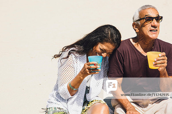 Senior couple sitting on beach  laughing  holding cold drinks  Long Beach  California  USA