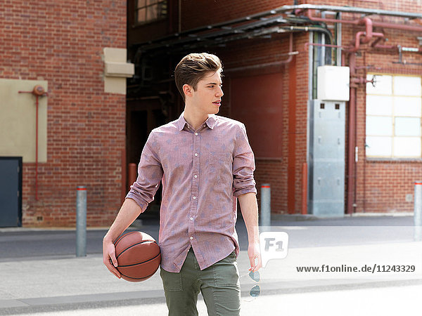 Junger Mann im Freien  hält Basketball und Sonnenbrille in der Hand und schaut weg
