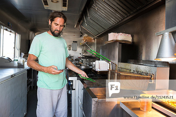 Mann kocht Essen im Fast-Food-Anhänger und sieht dabei auf ein Smartphone
