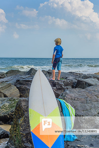Junge steht auf Felsen am Meer  schaut in die Ansicht  Surfbrett im Vordergrund