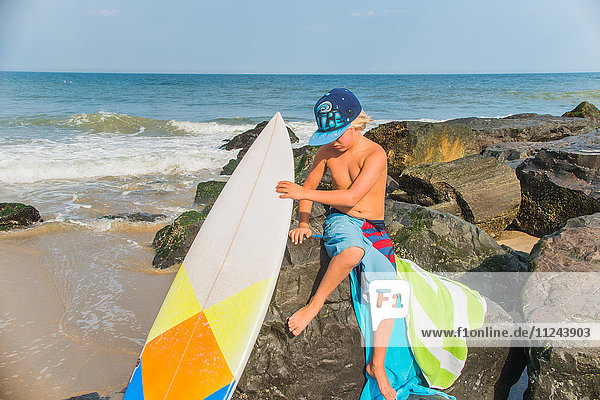 Junge sitzt am Strand auf Felsen und hält ein Surfbrett