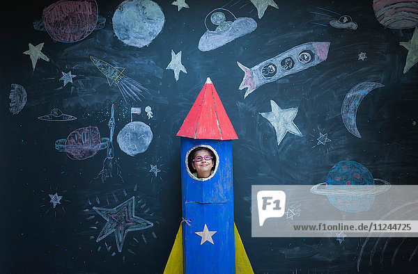 Portrait eines Mädchens in einer handgefertigten Weltraumrakete vor Kreidezeichnungen zum Thema Weltraum