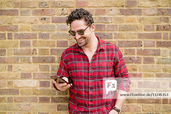 Young man leaning against brick wall reading smartphone texts