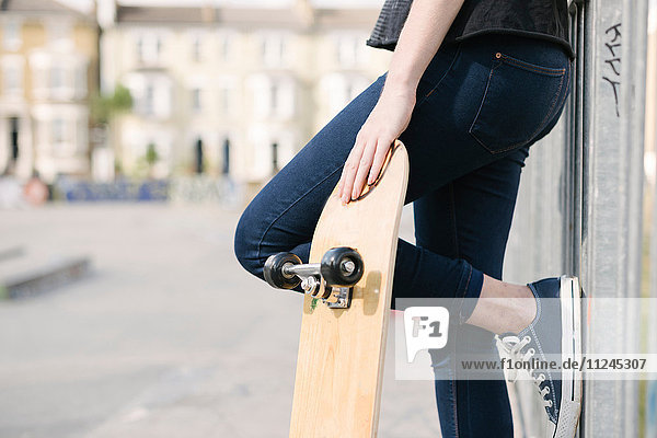Taille einer Skateboardfahrerin  die sich im Skatepark gegen einen Zaun lehnt