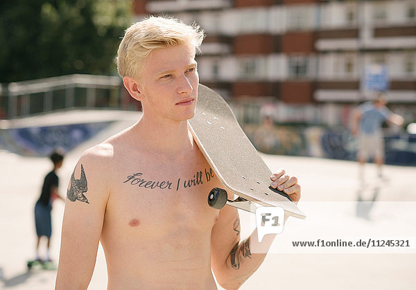 Tätowierter junger Mann mit Skateboard auf der Schulter im Skatepark