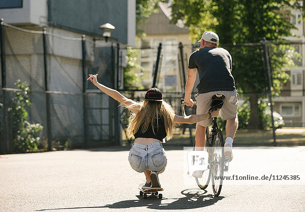 Rückansicht einer Skateboardfahrerin und eines Radfahrers auf dem Basketballplatz