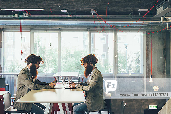Männliche erwachsene Hipster-Zwillinge sitzen von Angesicht zu Angesicht am Schreibtisch im Büro.