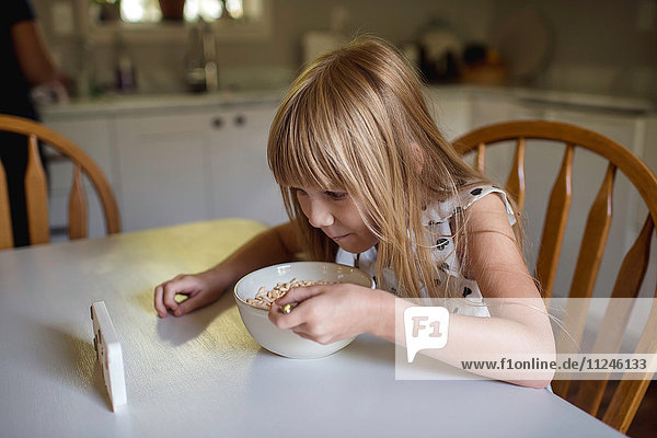 Kleines Mädchen beim Frühstücken  während es sich ein elektronisches Gerät ansieht.