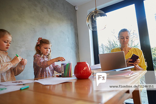 Mittlere erwachsene Frau liest Smartphone-Texte am Esstisch  während die Töchter malen