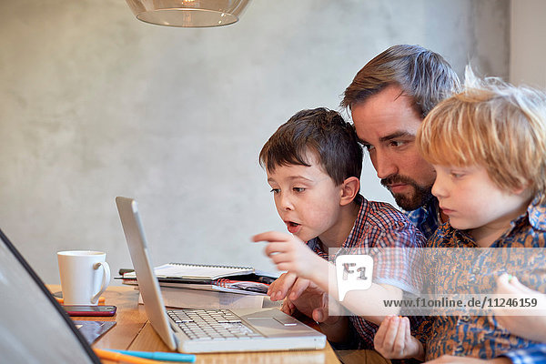 Mittelgroßer erwachsener Mann am Laptop mit zwei Söhnen am Esstisch