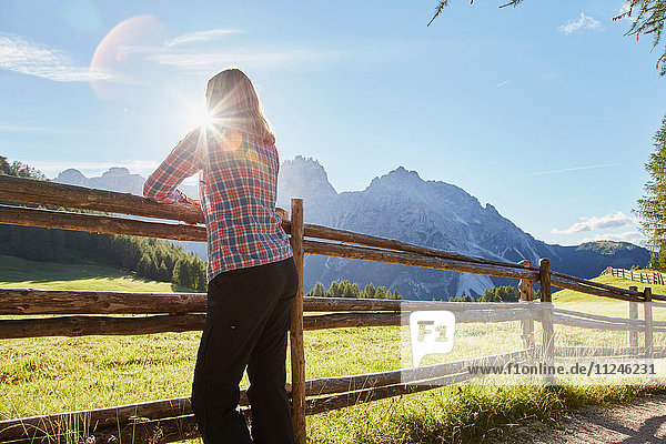 Reife Frau lehnt am Zaun und schaut auf die sonnenbeschienenen Dolomiten  Sexten  Südtirol  Italien