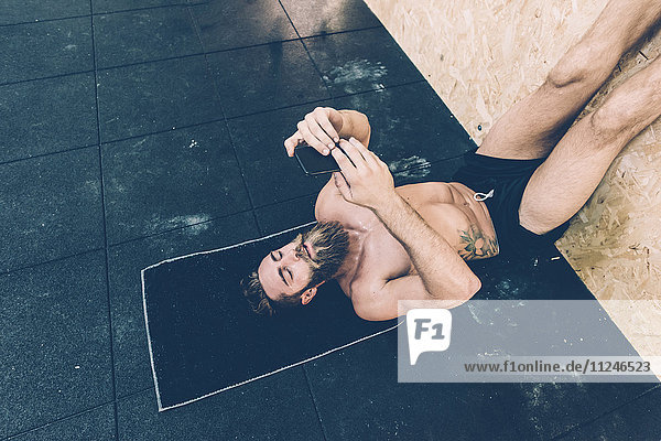 Männlicher Crosstrainer liest im Fitnessstudio auf dem Rücken liegend ein Smartphone
