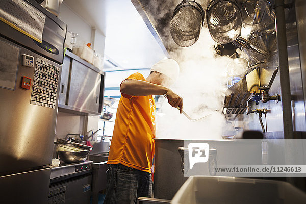 The ramen noodle shop kitchen. A vast pot on the stove  and steam rising. A man cooking noodles..