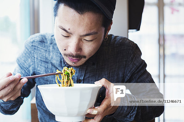 A ramen noodle cafe in a city. A man seated eating ramen noodles from a large broth bowl.