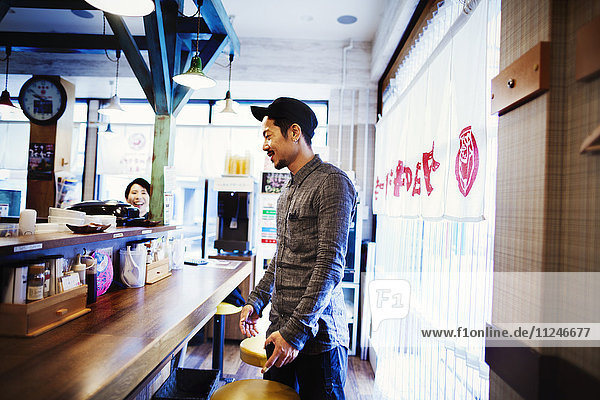 A ramen noodle cafe in a city. Customer standing up at the counter ordering food.