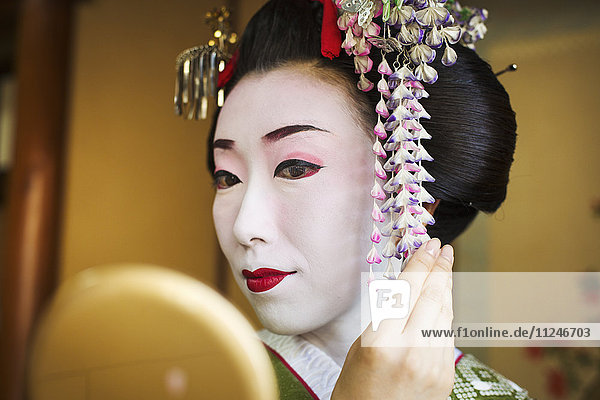 Eine Frau  geschminkt im traditionellen Geisha-Stil  mit aufwändiger Frisur und blumigen Haarspangen  mit weißem Gesichtsschminke mit leuchtend roten Lippen und dunklen Augen. Sie schaut in einen Handspiegel.