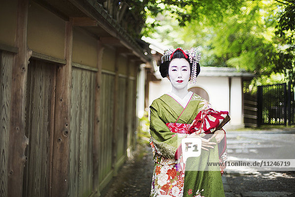 Eine im traditionellen Geisha-Stil gekleidete Frau in Kimono und Obi  mit aufwändiger Frisur und blumigen Haarspangen  mit weißer Gesichtsschminke mit leuchtend roten Lippen und dunklen Augen auf einer Straße.
