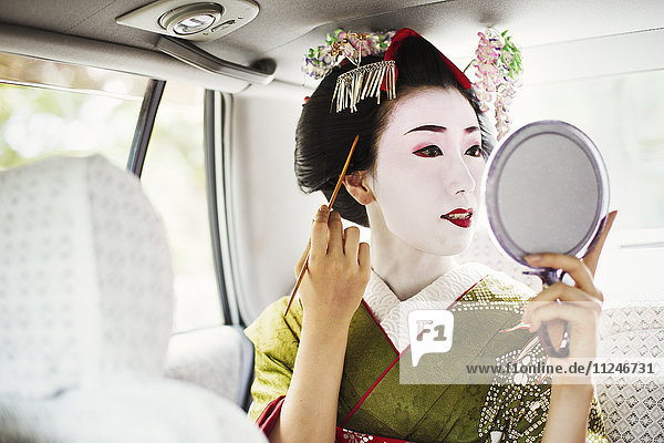 Eine im traditionellen Geisha-Stil gekleidete Frau in Kimono und Obi  mit aufwändiger Frisur und blumigen Haarspangen  mit weißer Gesichtsschminke mit leuchtend roten Lippen und dunklen Augen in einem Auto mit einem Handspiegel.