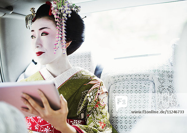 Eine im traditionellen Geisha-Stil gekleidete Frau  in einem Kimono mit aufwändiger Frisur und blumigen Haarspangen  mit weißem Gesichtsschminke  die ein digitales Tablett hält  reist in einem Auto.