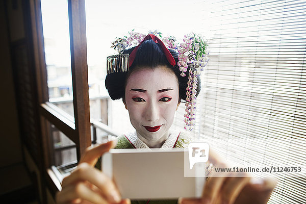 A woman dressed in the traditional geisha style  wearing a kimono and obi  with an elaborate hairstyle and floral hair clips  with white face makeup with bright red lips and dark eyes taking a selfie of herself.