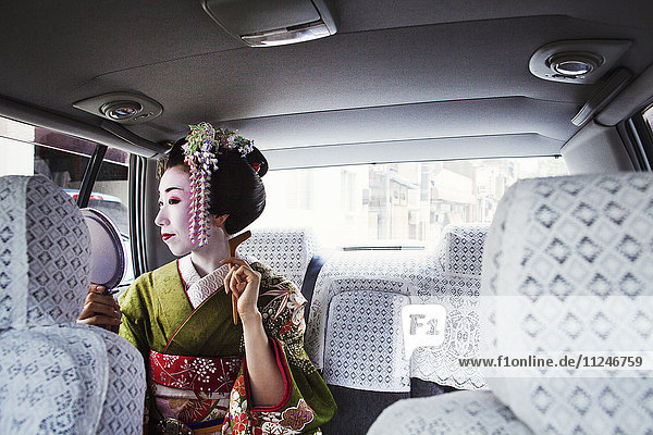 Eine im traditionellen Geisha-Stil gekleidete Frau in einem Kimono mit aufwändiger Frisur und blumigen Haarspangen  mit weißer Gesichtsschminke mit leuchtend roten Lippen und dunklen Augen in einem Auto mit einem Handspiegel.