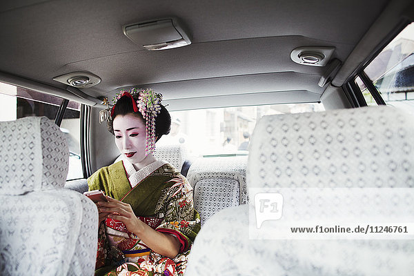 Eine im traditionellen Geisha-Stil gekleidete Frau in einem Kimono mit aufwändiger Frisur und blumigen Haarspangen  mit weißem Gesichts-Make-up mit leuchtend roten Lippen und dunklen Augen in einem Auto mit einem Smartphone